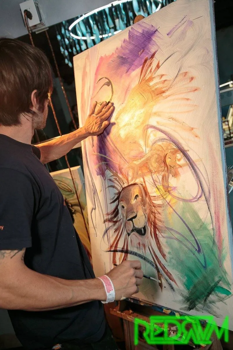 A person is painting a vibrant image of a lion on a canvas using their hands and brushes. Bright colors and dynamic strokes are visible.
