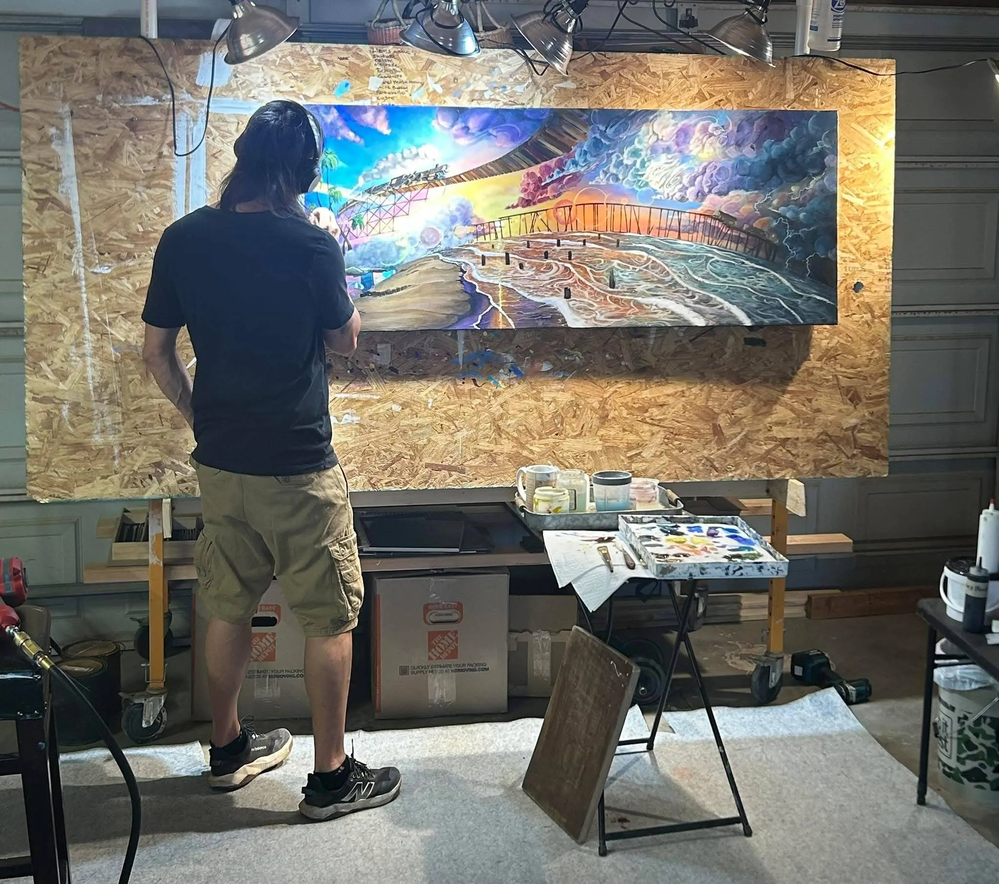 A person in a black shirt and khaki shorts is painting a colorful landscape on a large canvas in a garage studio. Art supplies and tools are visible on tables nearby.